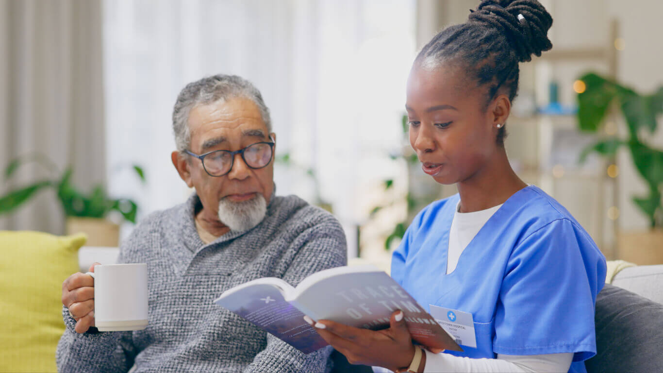A female nurse is reading a book to a senior man during hospice care at home.