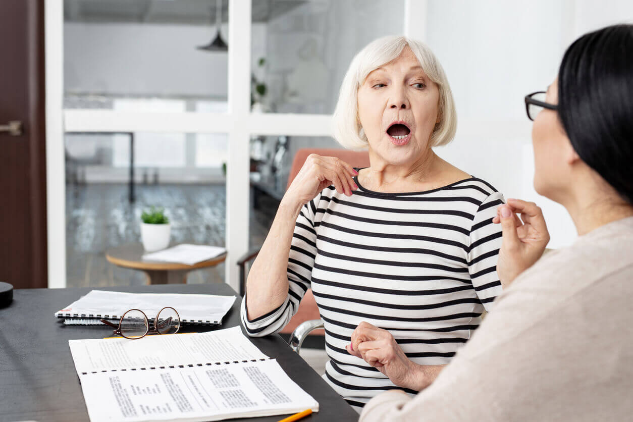 A focused female senior tutor demonstrates pronunciation during in-home speech therapy.