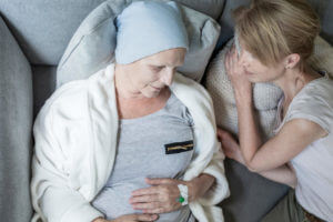 A female caregiver and an ill senior woman lying in bed together during end-of-life care at home for the patient undergoing cancer treatment.