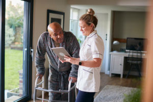 A senior man in a dressing gown is using a walking frame and being assisted by a female nurse with a digital tablet during home health services.