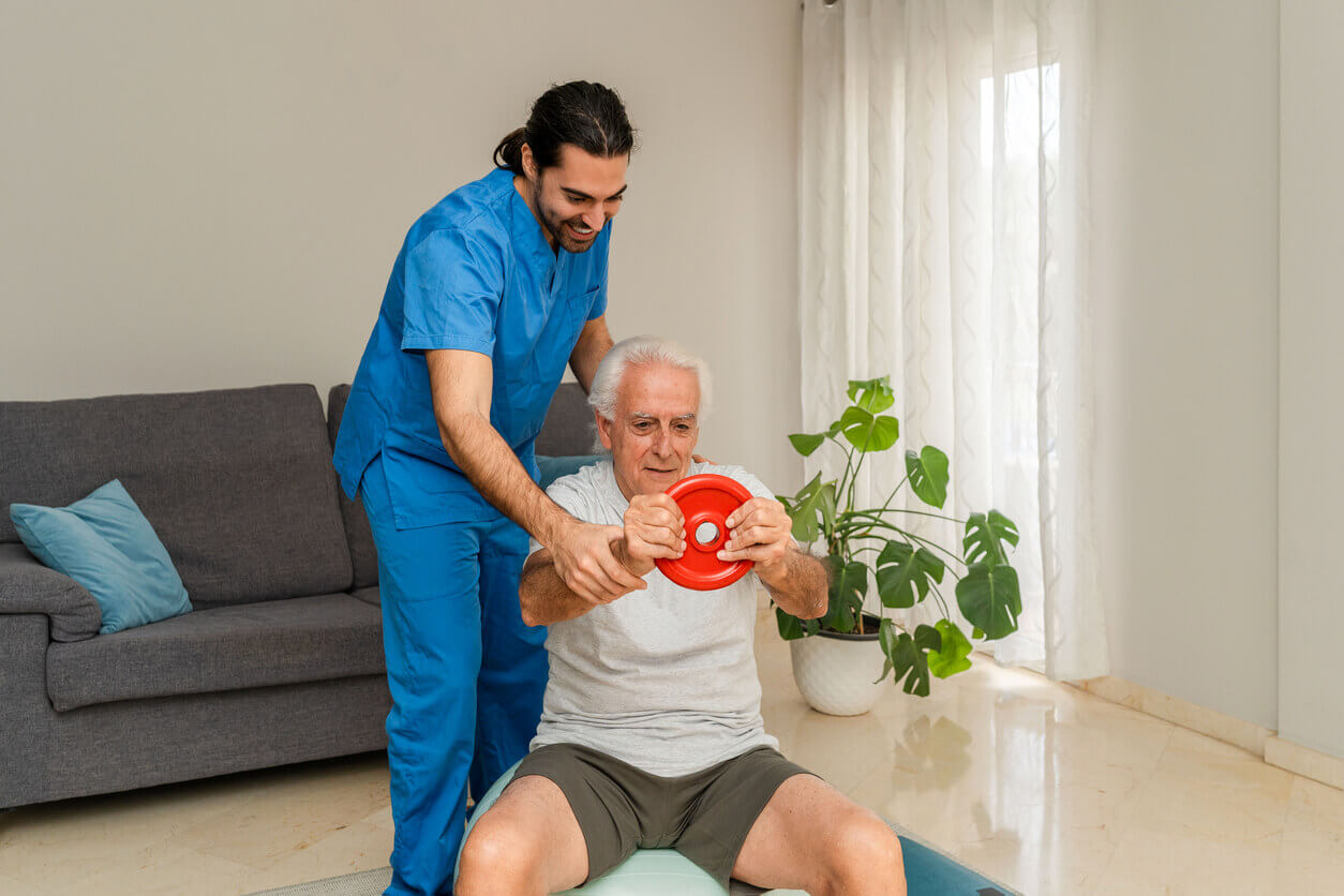 A male physical therapist in a blue uniform assists a senior man in exercising with weights during in-house physical rehabilitation services.