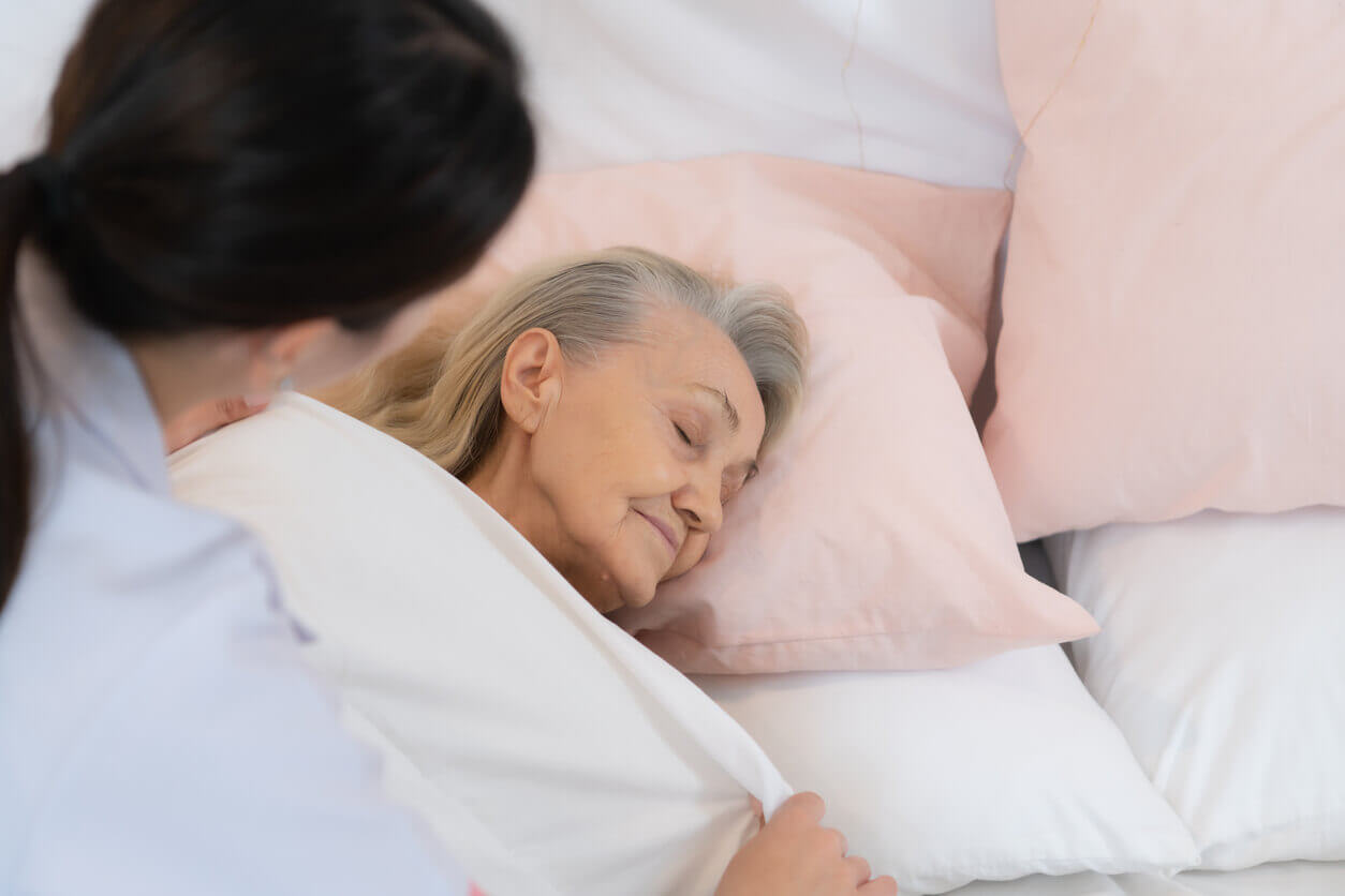 A hospice caregiver gently covers an elderly woman with dementia as she rests in bed, offering comfort and warmth.